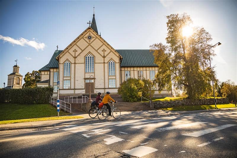 Kuvassa kirkko, tie, aurinko ja kaksi polkupyöräilijää.